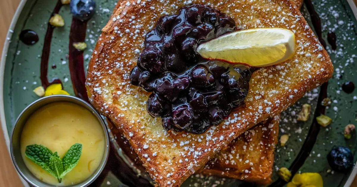 Delicious French toast topped with blueberry compote, a lemon wedge, and powdered sugar, served with a side of lemon curd garnished with fresh mint.