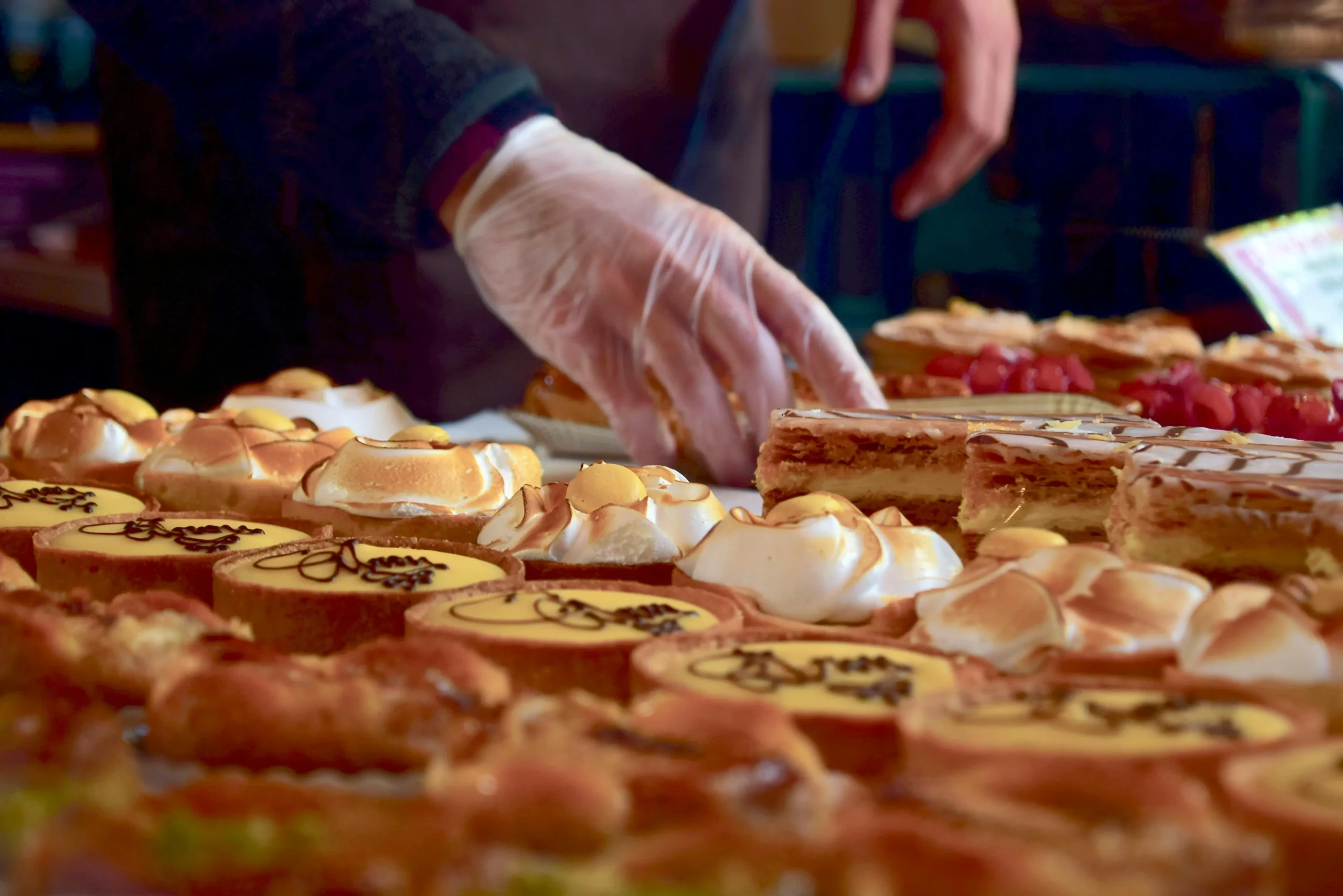 A pastry chef wearing gloves arranges an assortment of freshly baked pastries, including lemon tarts, mille-feuille, and meringue-topped treats, at a bakery display.